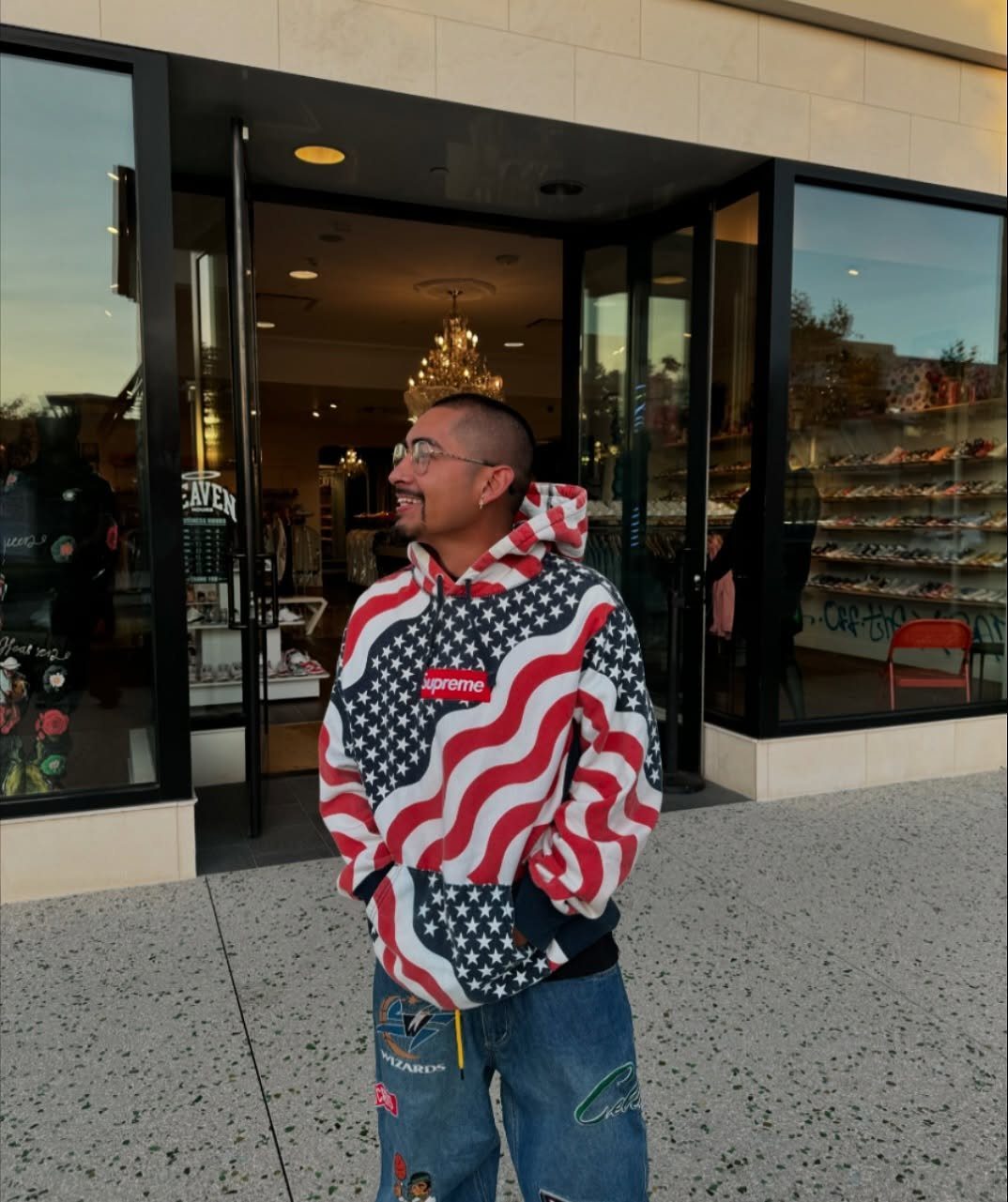 Man wearing a Supreme patterned hoodie and patched denim jeans standing outside a store.
