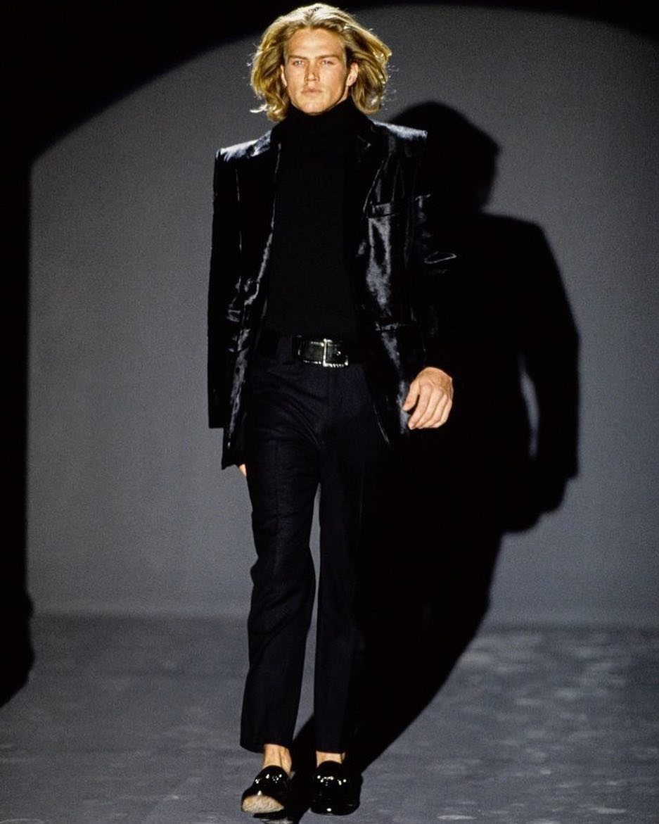 A male model walking a runway in sleek black evening wear and a blazer.