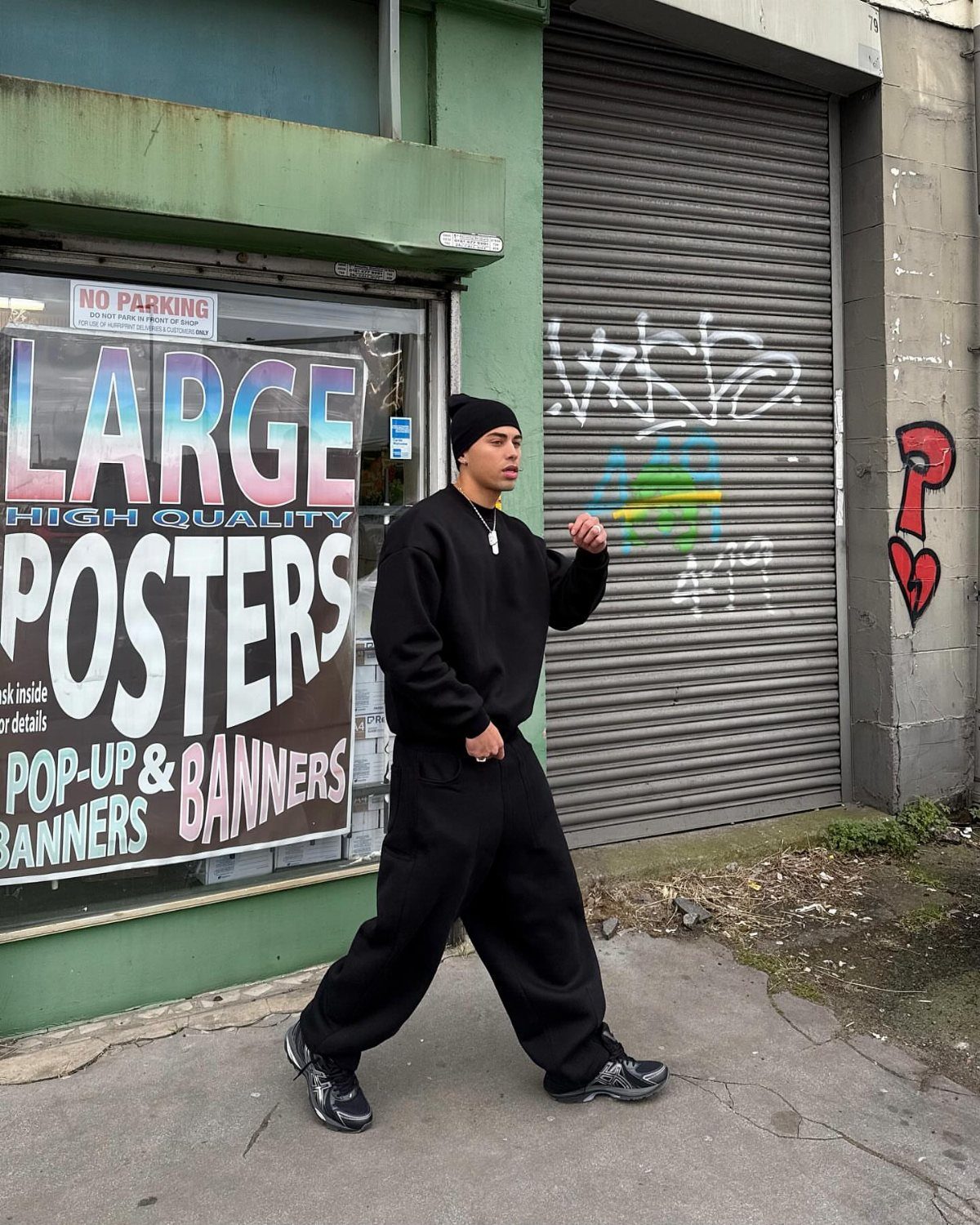 Man in an all-black streetwear outfit walking past a storefront with large posters.