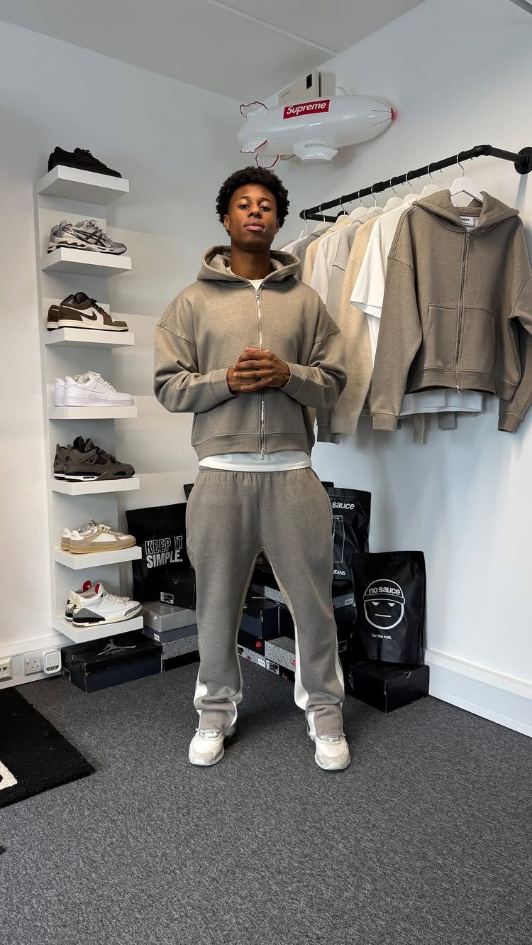 A man in a grey tracksuit and white sneakers standing in a room filled with clothing and shoes.