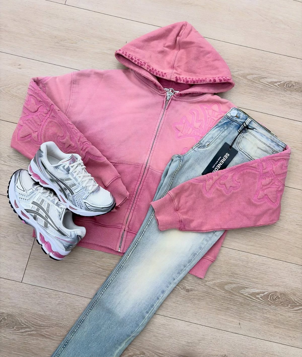 Pink hoodie, light blue jeans, and silver Asics sneakers arranged on a wooden floor.
