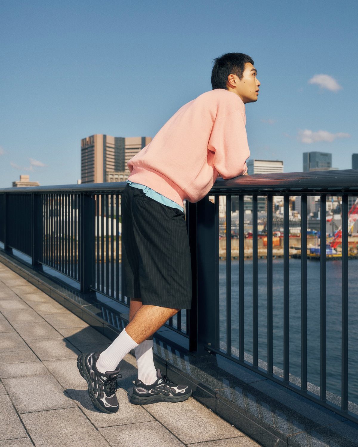 Man wearing a pink sweatshirt and black shorts leaning on a railing by the water.
