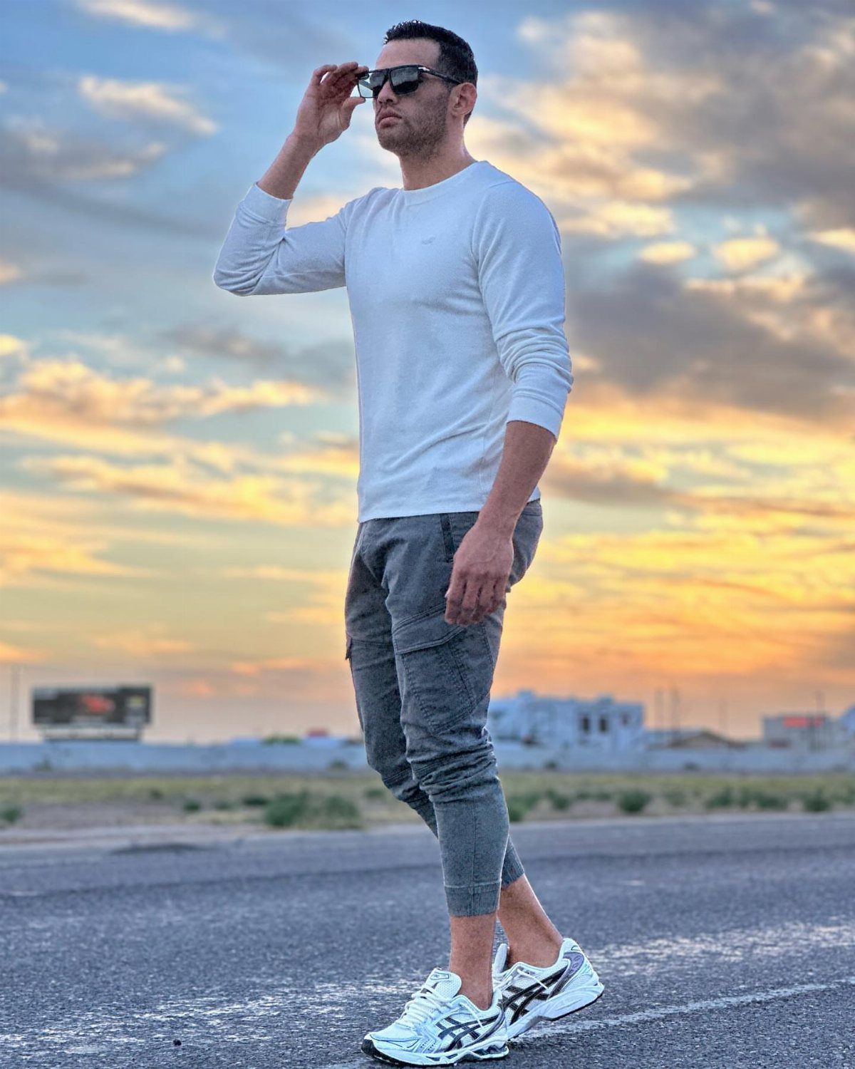 Man in white long-sleeve shirt and grey trousers wearing white athletic sneakers at sunset.