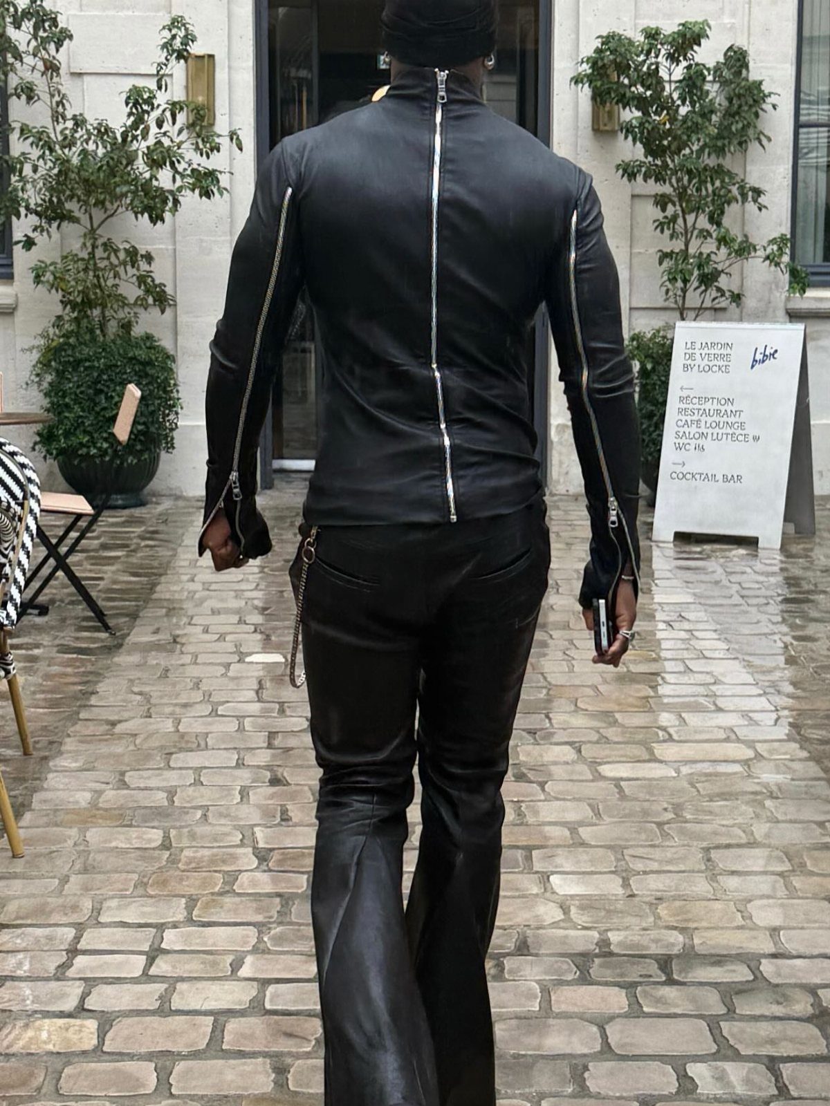 A person in tight black leather clothing walking through a cobblestone courtyard towards a building.