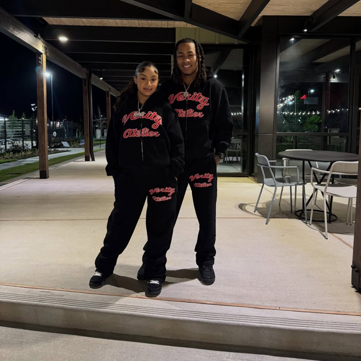 A man and woman posing in matching black graphic hoodies and sweatpants at night.