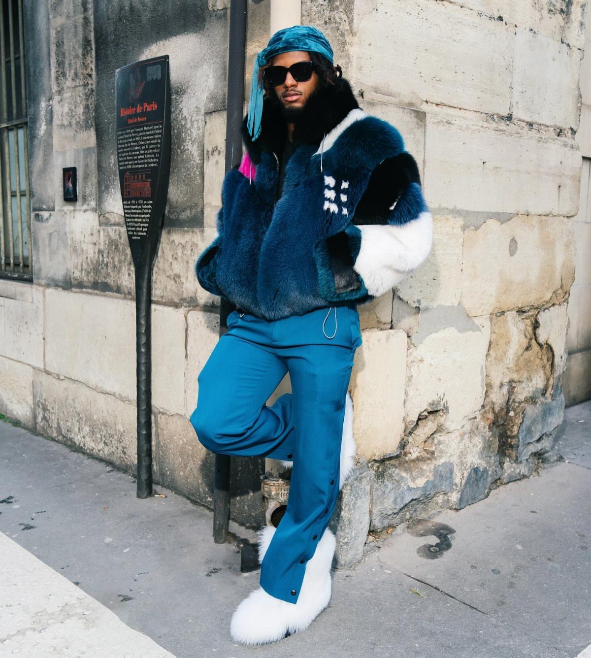 Man in voluminous blue faux fur jacket and matching trousers posing against a stone wall.