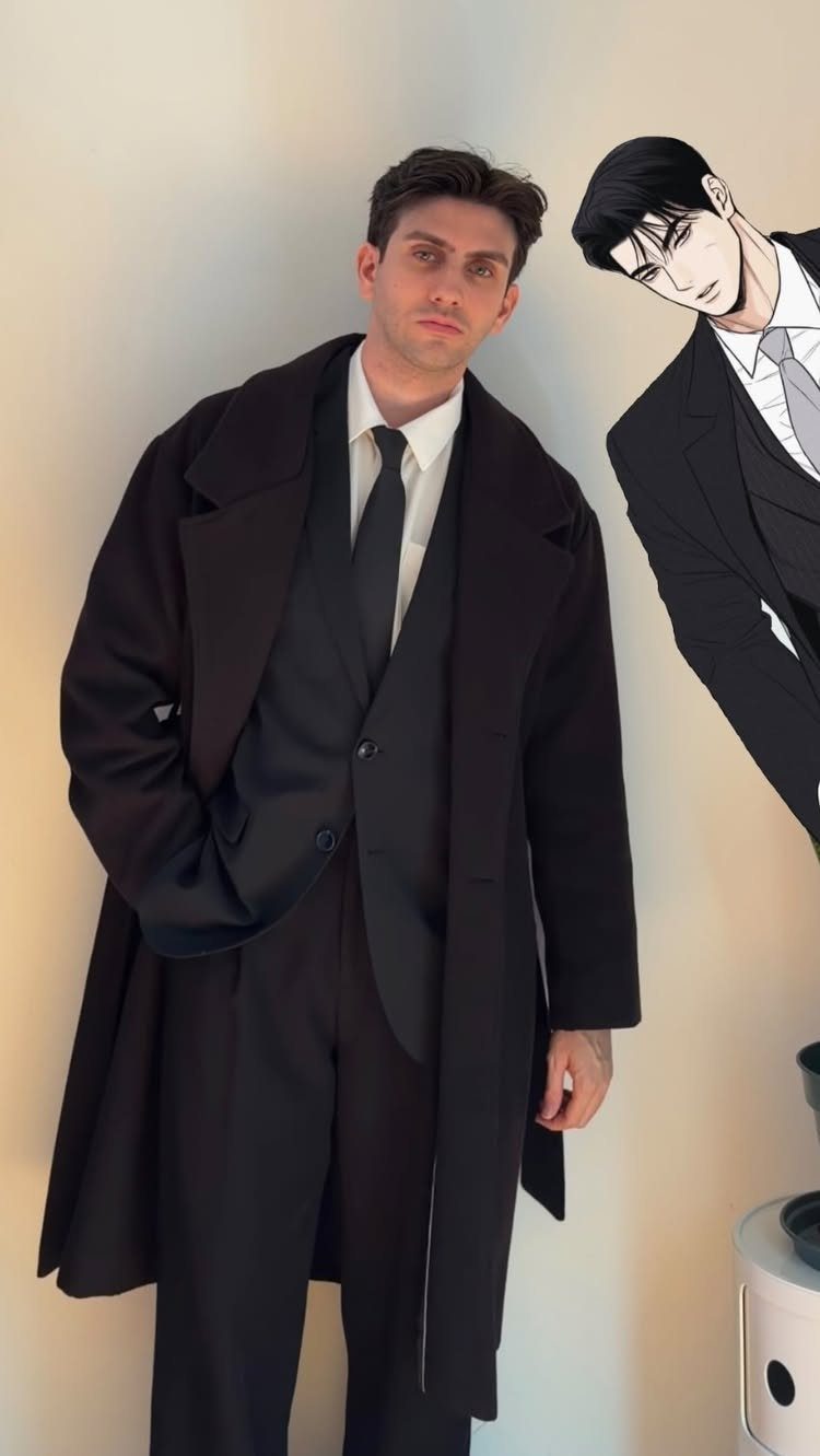 Man in a black three-piece suit and long overcoat standing next to an anime cutout.