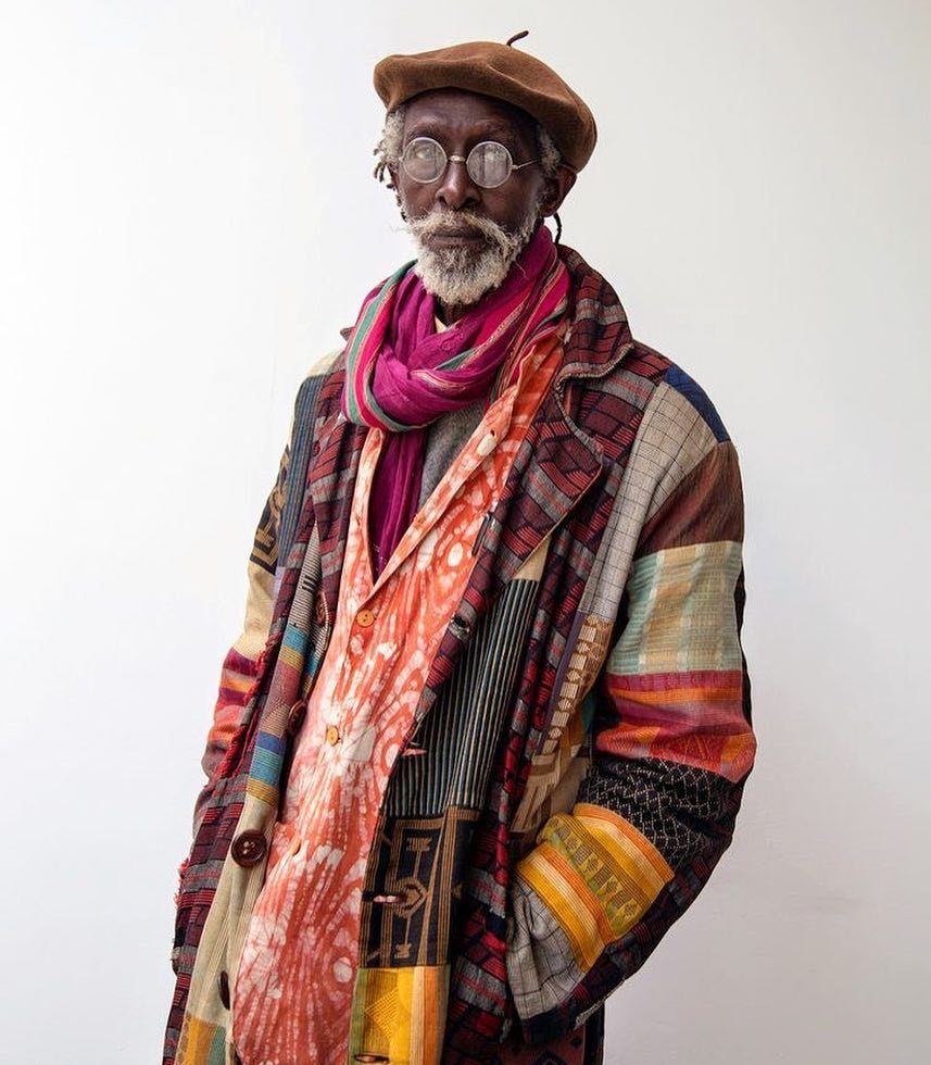 An older man with a white beard wearing a colorful patchwork coat, pink scarf, and brown beret.