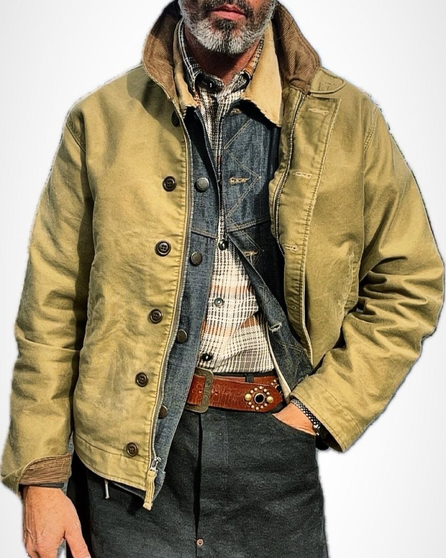 Man wearing a tan canvas jacket over a denim vest and plaid shirt.