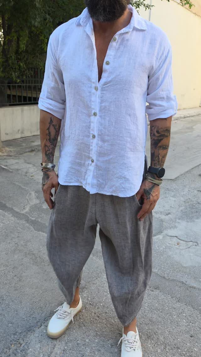 Man wearing a white linen shirt and loose grey trousers with tattoos on his arms.
