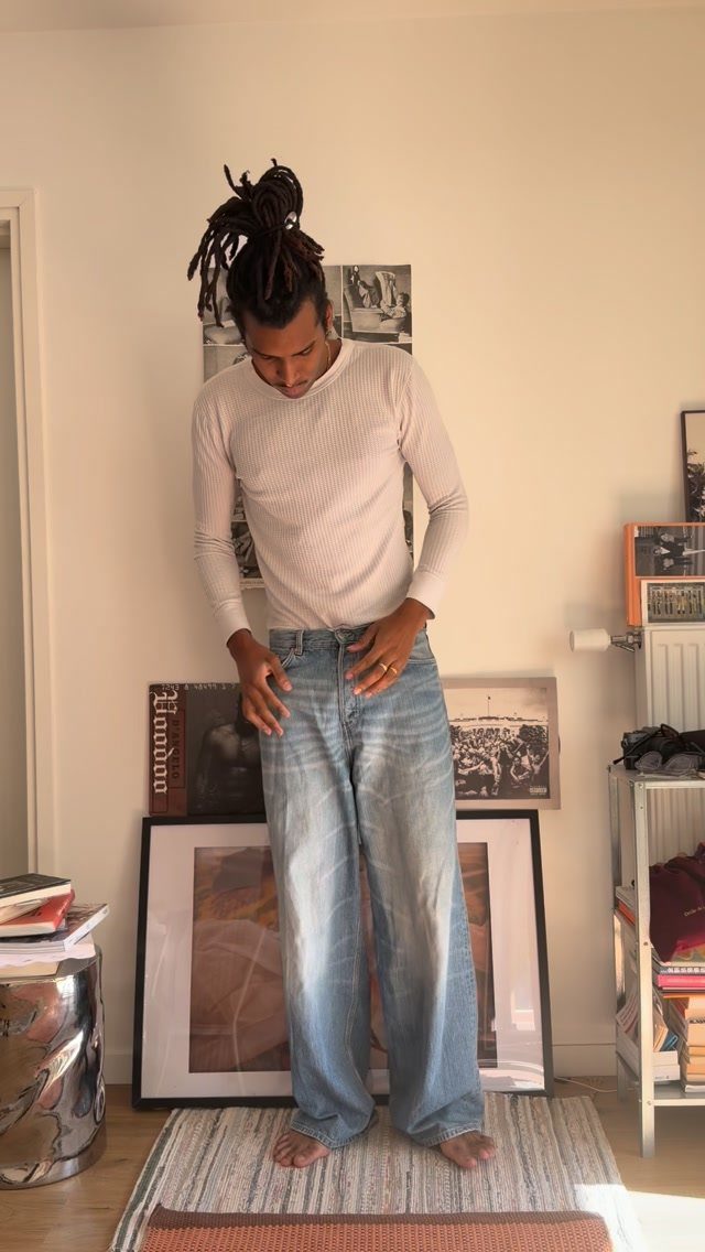 A man with dreadlocks wearing a ribbed white shirt and wide-leg blue jeans.