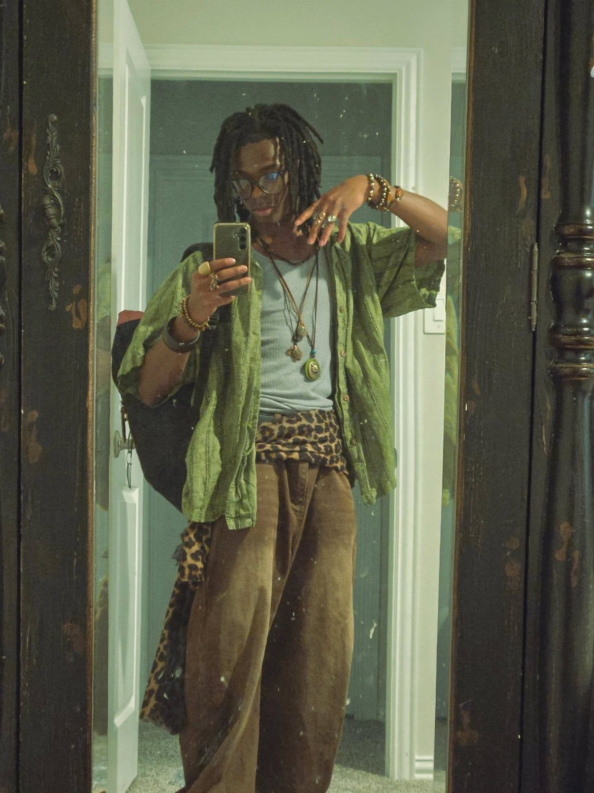 A man with dreadlocks posing in a mirror wearing green boho-style clothing and layered jewelry.