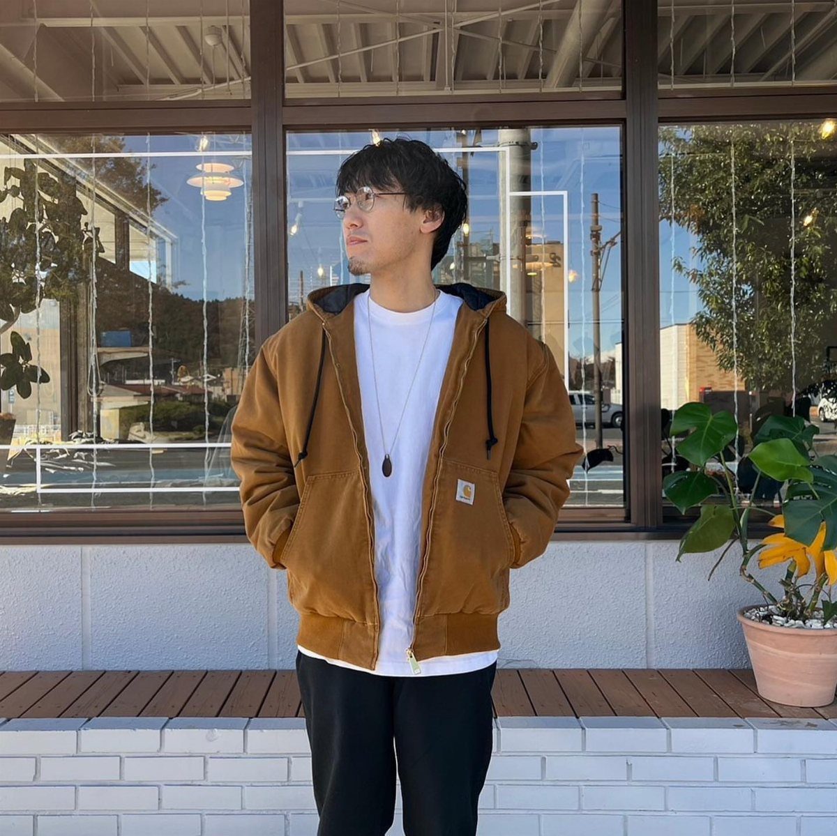 Man wearing a brown hooded jacket and white t-shirt standing in front of a window.