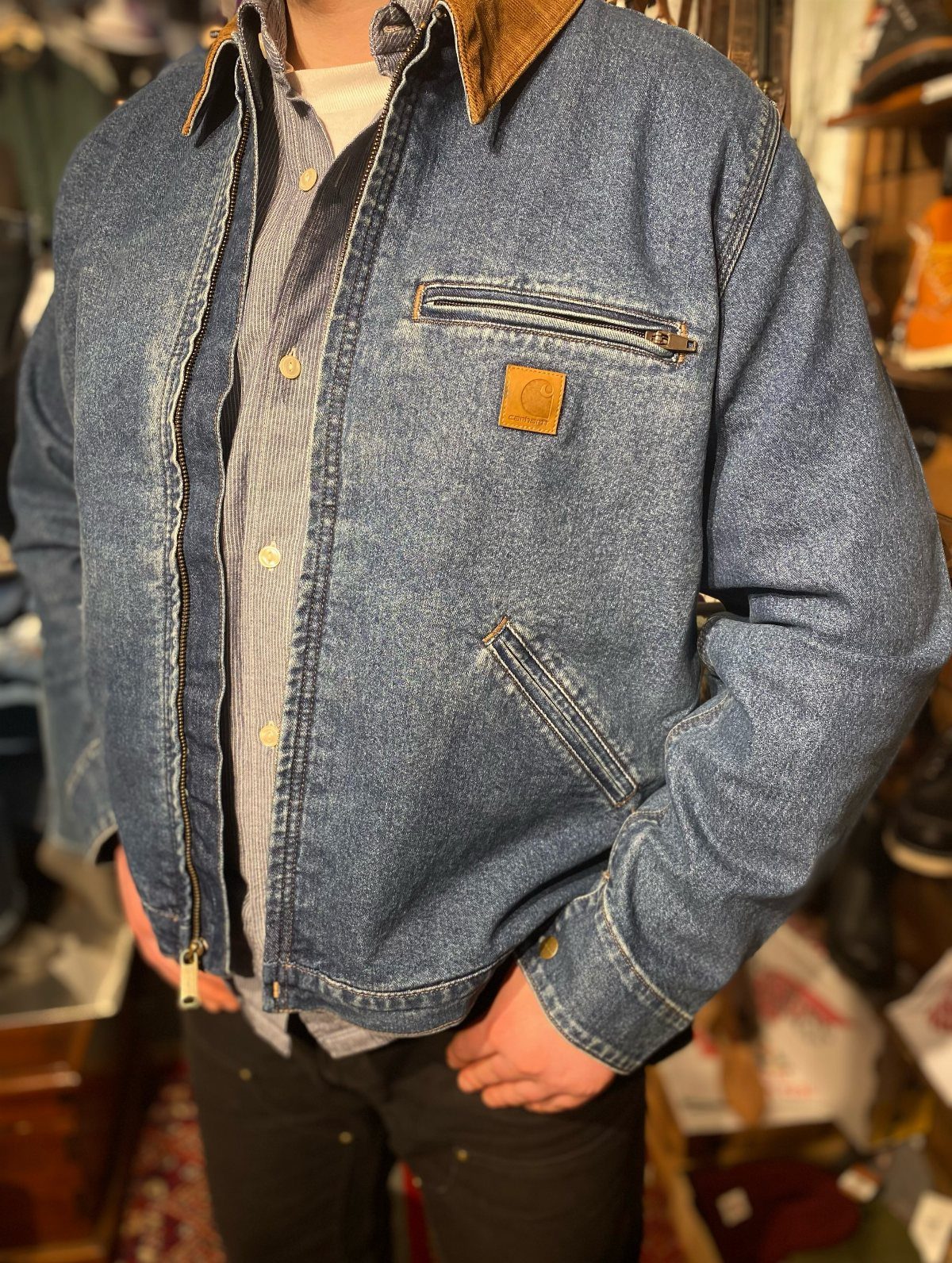 A person wearing a blue denim Carhartt jacket with a brown corduroy collar and black pants.