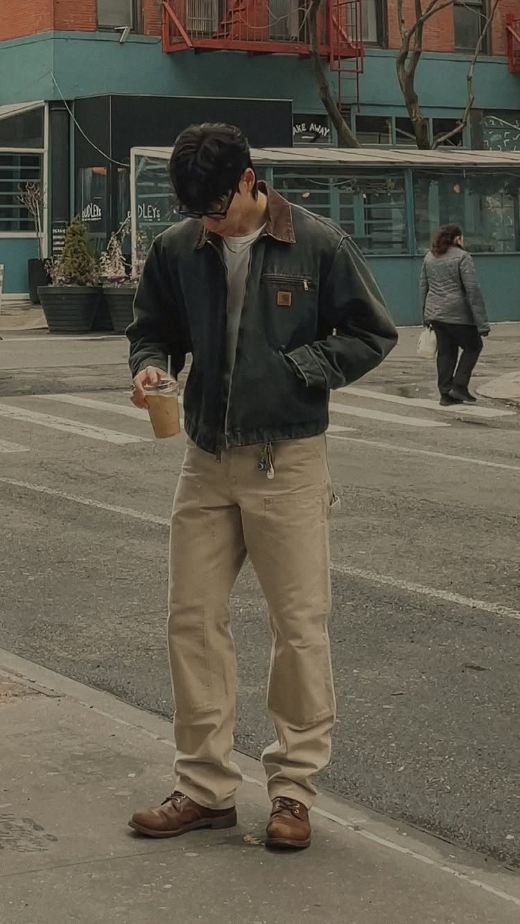 Man in a dark green Carhartt jacket, tan pants, and brown boots standing on a city street.