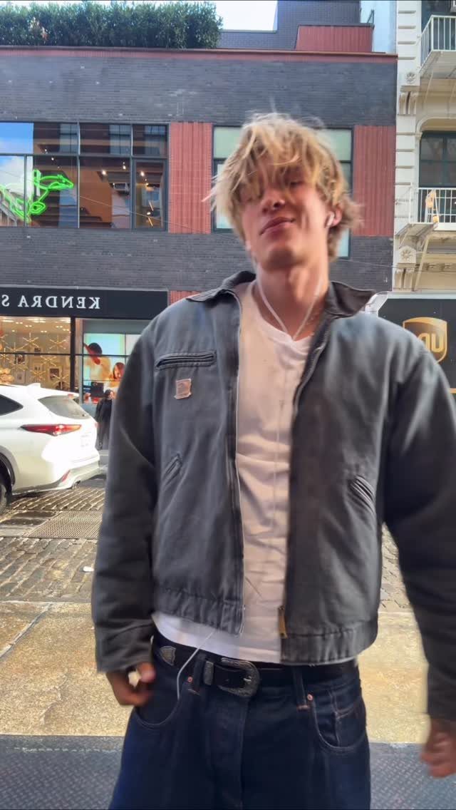Young man with blonde hair wearing a grey Carhartt jacket, white t-shirt, and dark blue jeans.