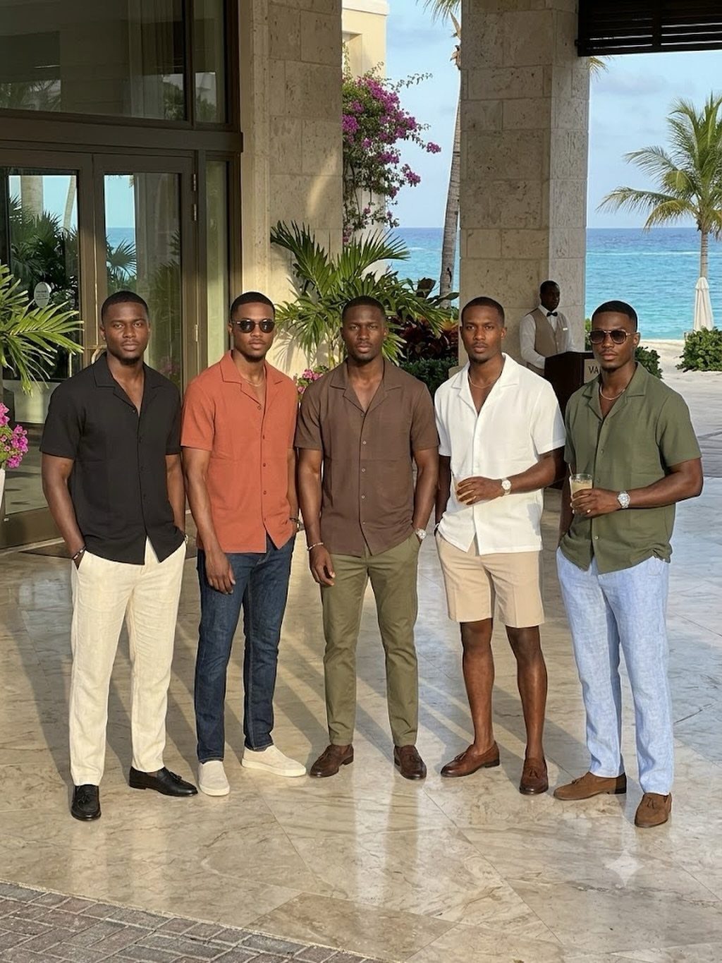 Five men posing in various colorful short-sleeved button-down shirts and light trousers near a tropical beach.