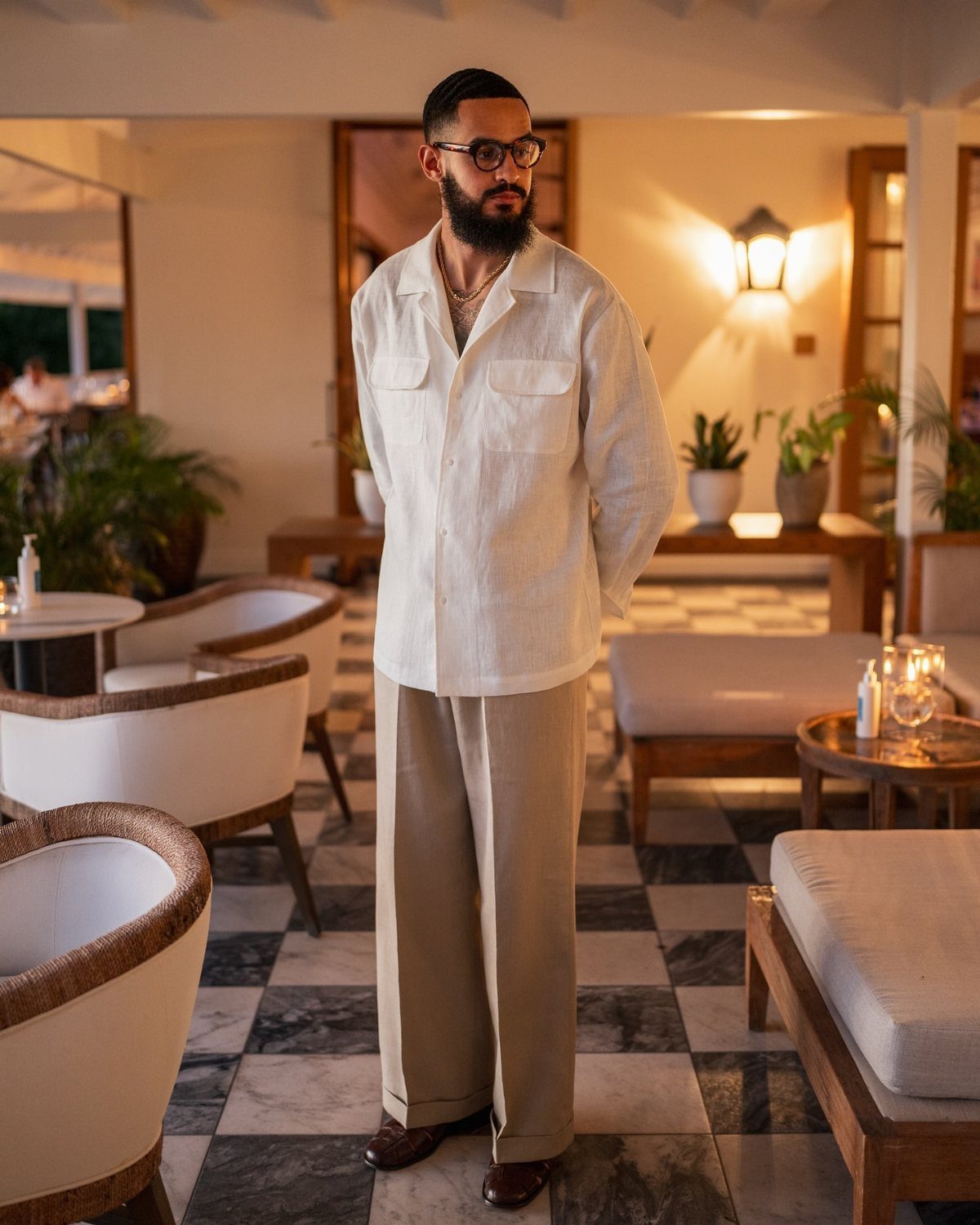 Man wearing cream linen shirt and beige wide-leg lightweight trousers standing in a lounge.