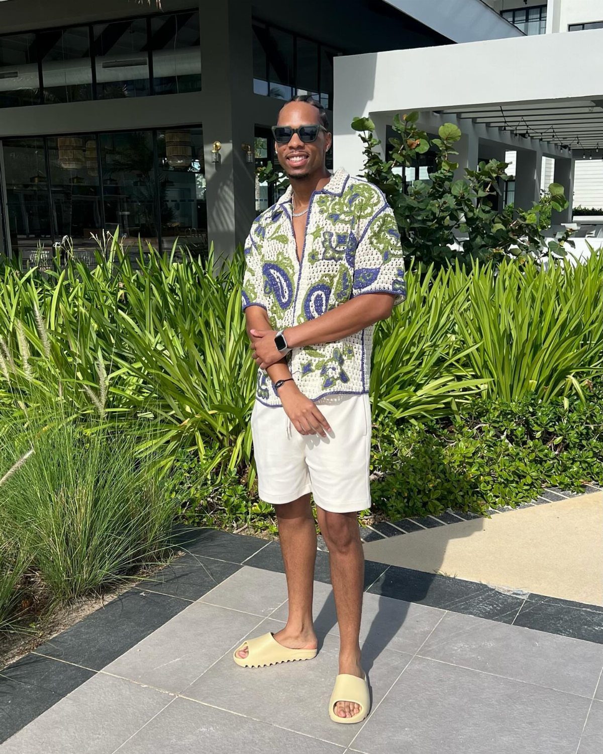 A man in a patterned tropical shirt and white shorts standing in front of green plants.
