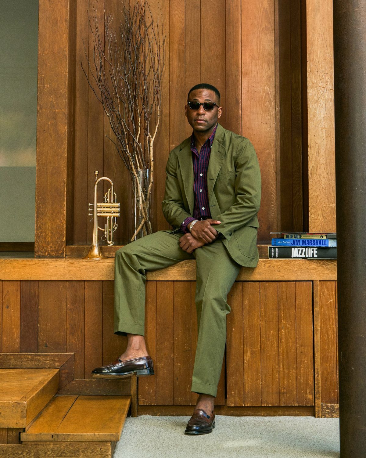 Man in olive green unstructured cotton jacket and matching trousers sitting on a wooden ledge.