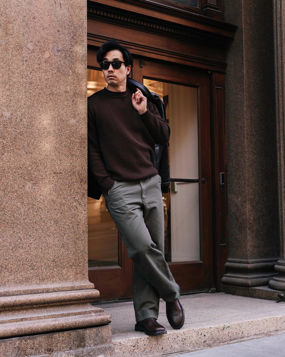 Man in a brown knit sweater, grey trousers, and dark brown Chelsea boots leaning against a stone pillar.