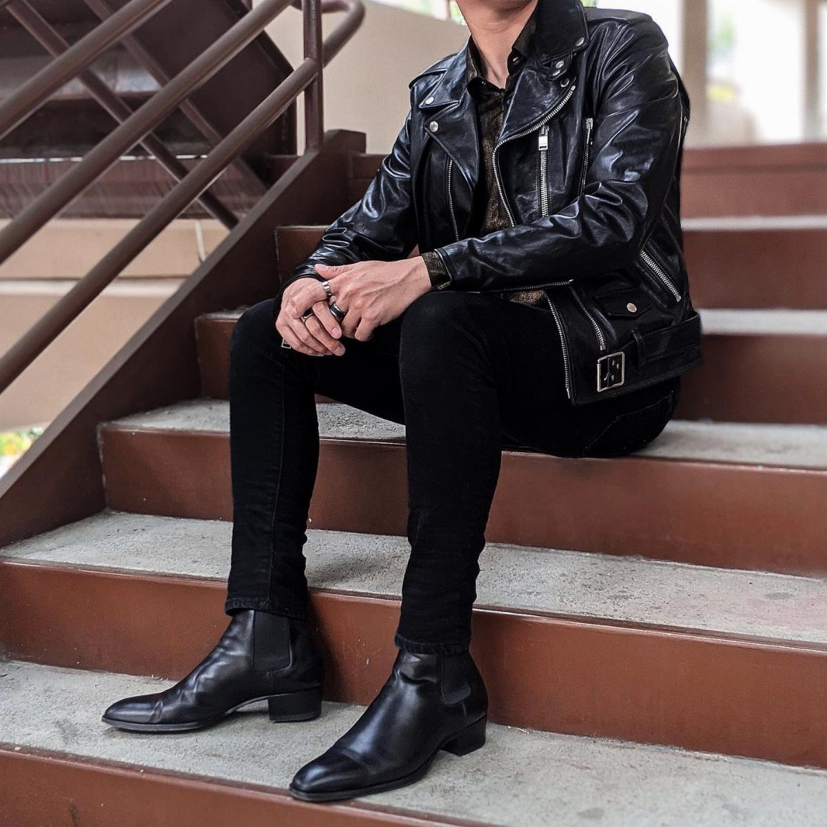 Man sitting on stairs wearing a black leather jacket, black jeans, and black Chelsea boots.