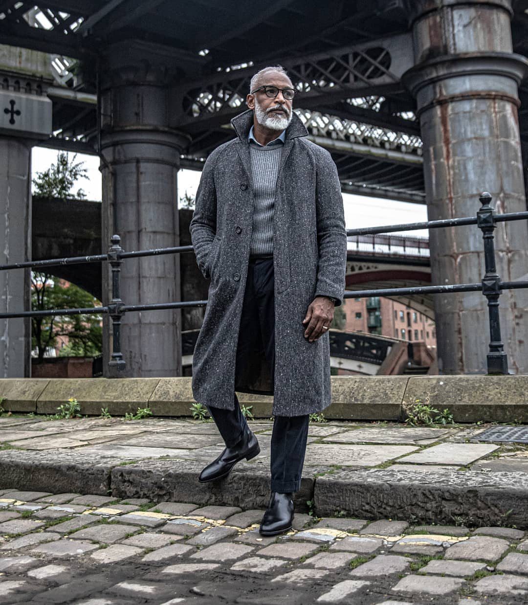 Man in a grey long overcoat and black Chelsea boots standing on cobblestones.