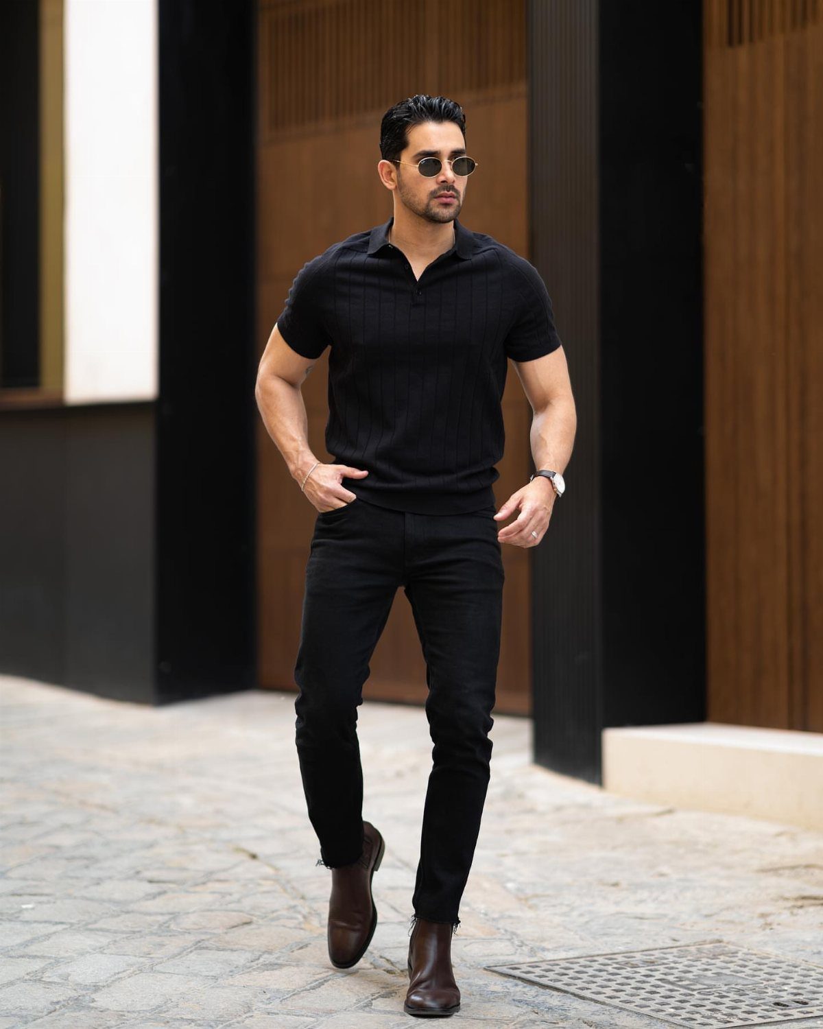 Man walking in a black polo shirt, black trousers, and brown leather Chelsea boots.