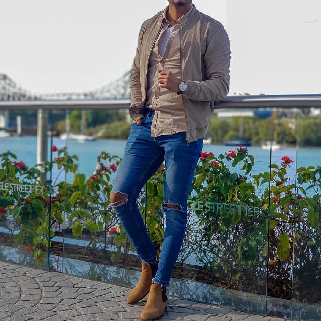Man wearing a tan jacket, blue ripped jeans, and tan suede Chelsea boots by a waterfront.