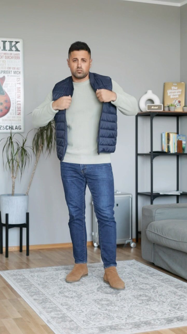 A man wearing a light grey sweater, navy vest, blue jeans, and tan suede Chelsea boots.