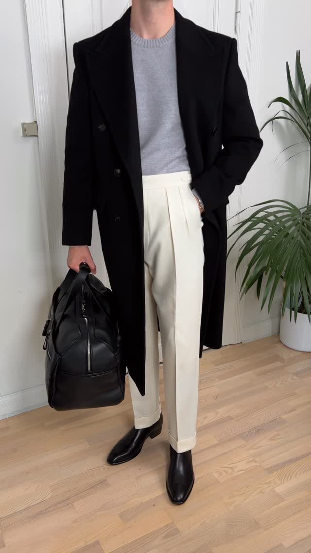 Man wearing a black overcoat, grey sweater, cream trousers, and black Chelsea boots.