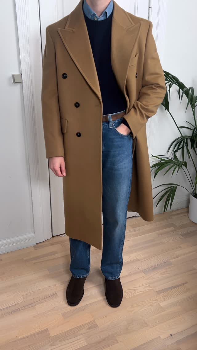 Man wearing a tan overcoat, navy sweater, blue jeans, and brown suede Chelsea boots standing indoors.