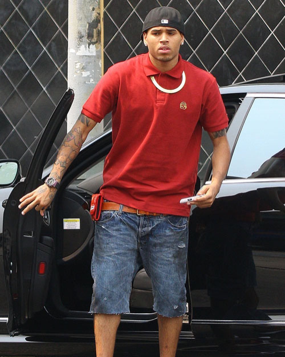Man in a red polo shirt and denim shorts standing by a black car.