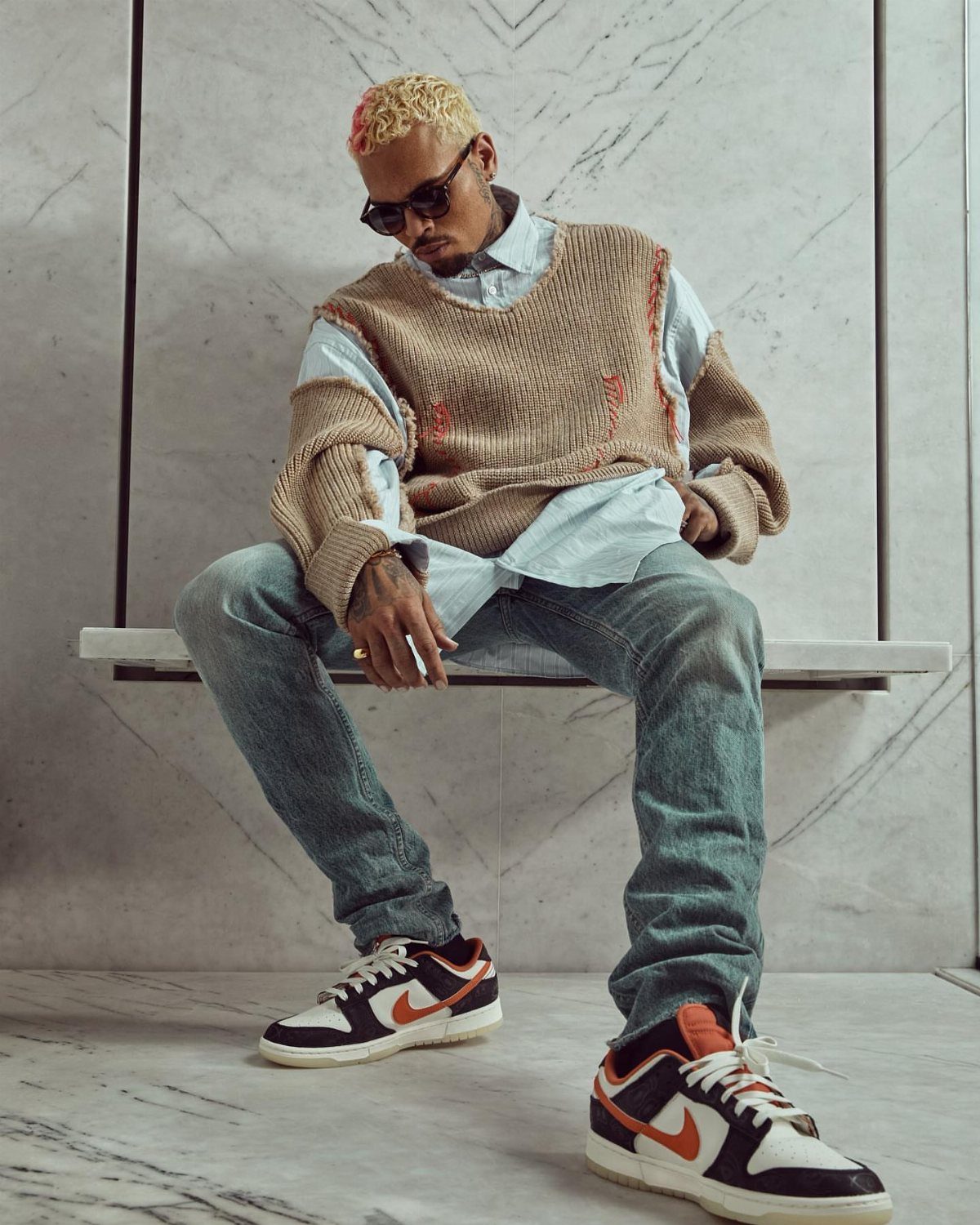 A man with blonde hair wearing a tan sweater, blue shirt, jeans, and sneakers sits on a ledge.