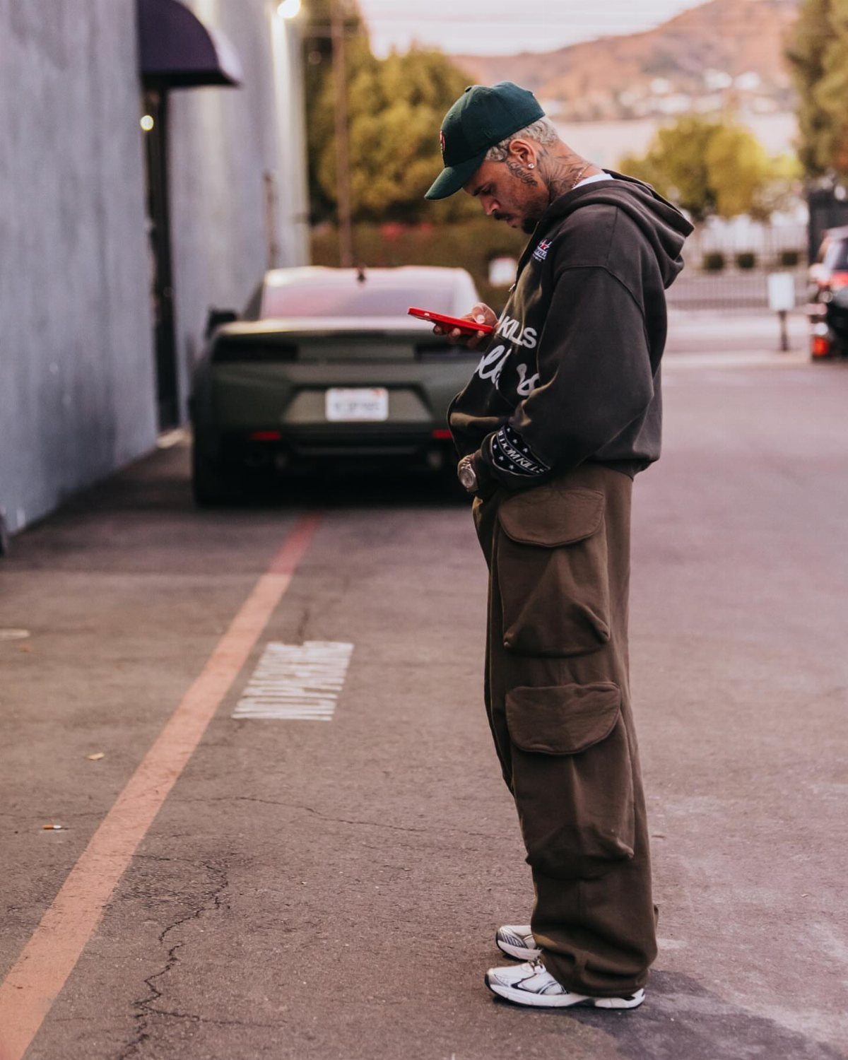 A man in a dark hoodie, green cap, and brown cargo pants looking at his phone.