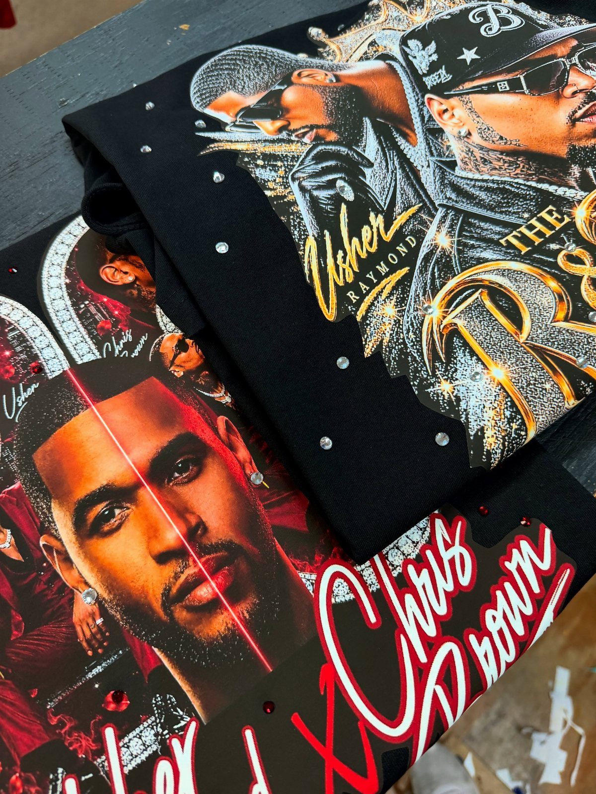 Two black graphic t-shirts featuring portraits of R&B singers Usher and Chris Brown.