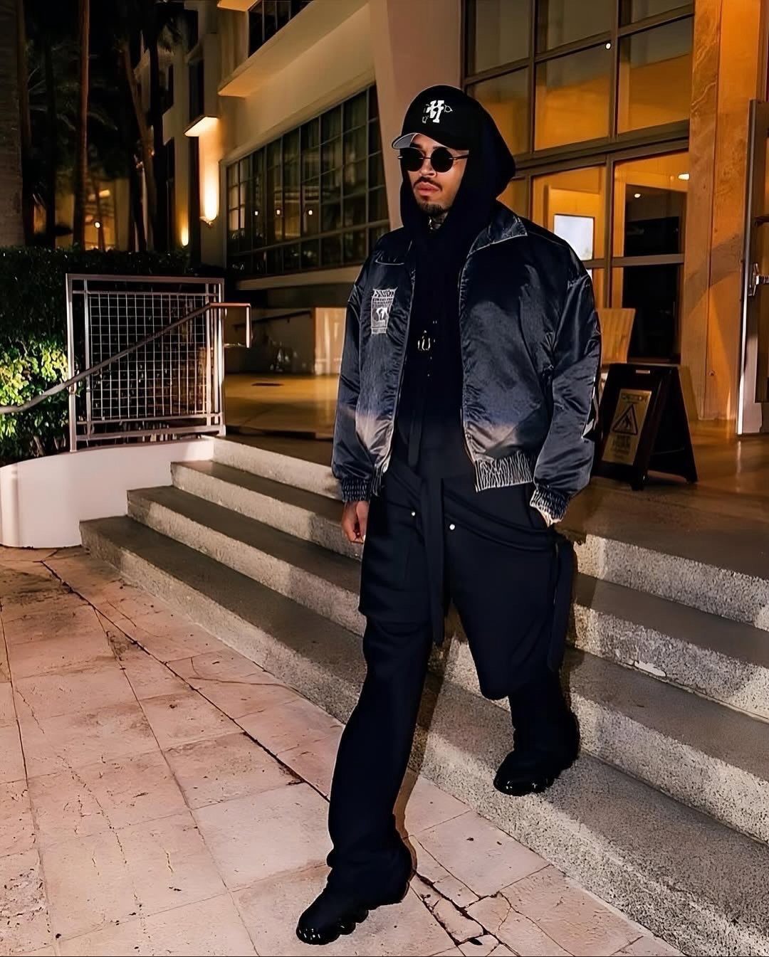 Man in urban streetwear posing on stairs wearing a bomber jacket, hoodie, and baseball cap.