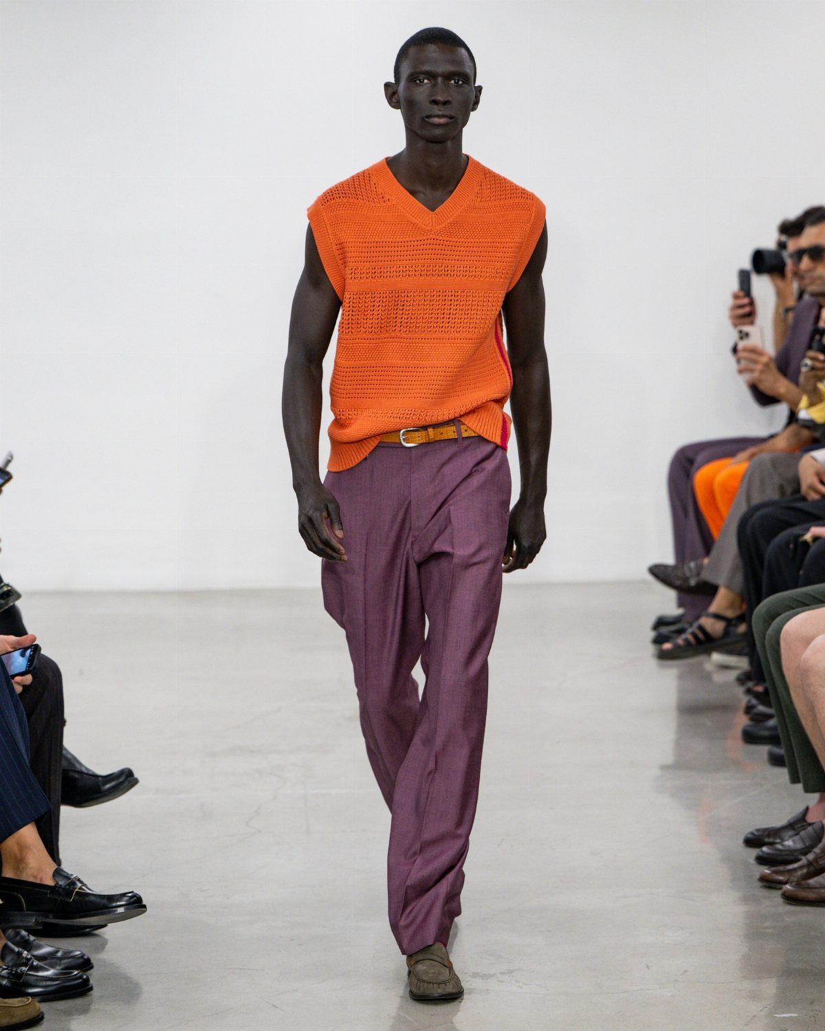 A male model walking a runway in an orange knit vest and purple trousers.