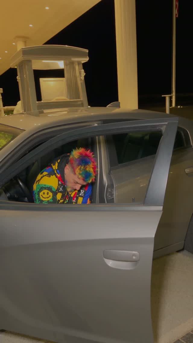 A person with rainbow hair wearing a colorful graphic jacket sitting inside a silver car.