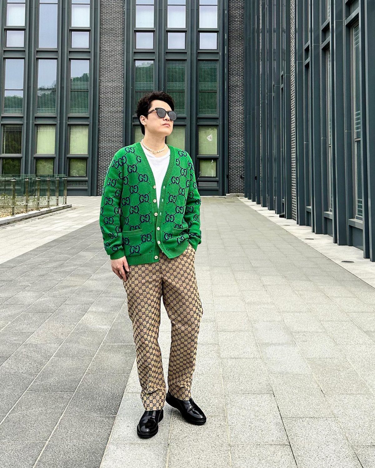 Man in green patterned cardigan and monogrammed tan trousers standing on a stone walkway.