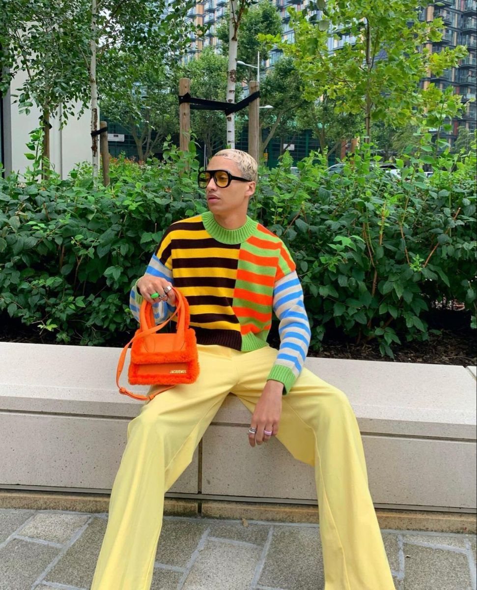 A person wearing a vibrant striped sweater and yellow pants sitting outdoors near green foliage.