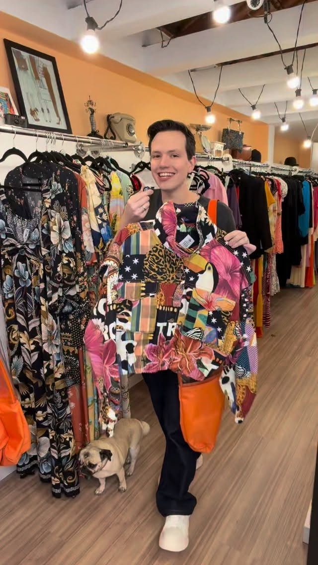 A smiling person holding up a brightly patterned garment in a clothing store with a pug.