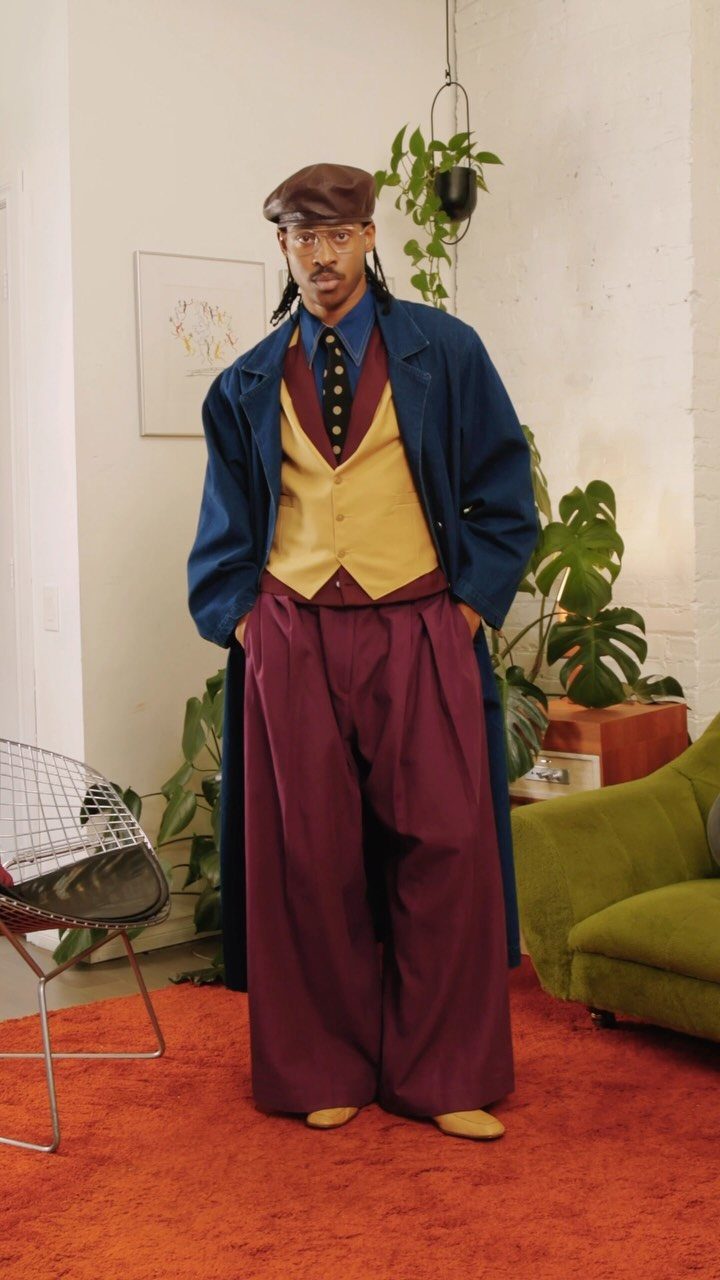Man wearing a colorful layered outfit including a blue coat, yellow vest, and maroon wide-leg trousers.