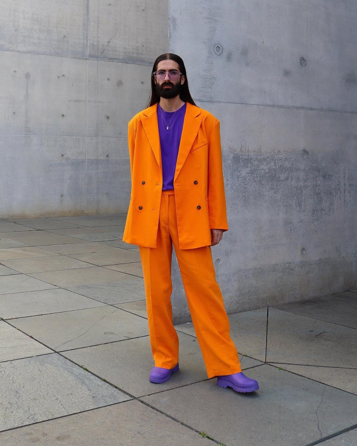 Man in a vibrant orange suit and purple t-shirt standing against a grey concrete wall.