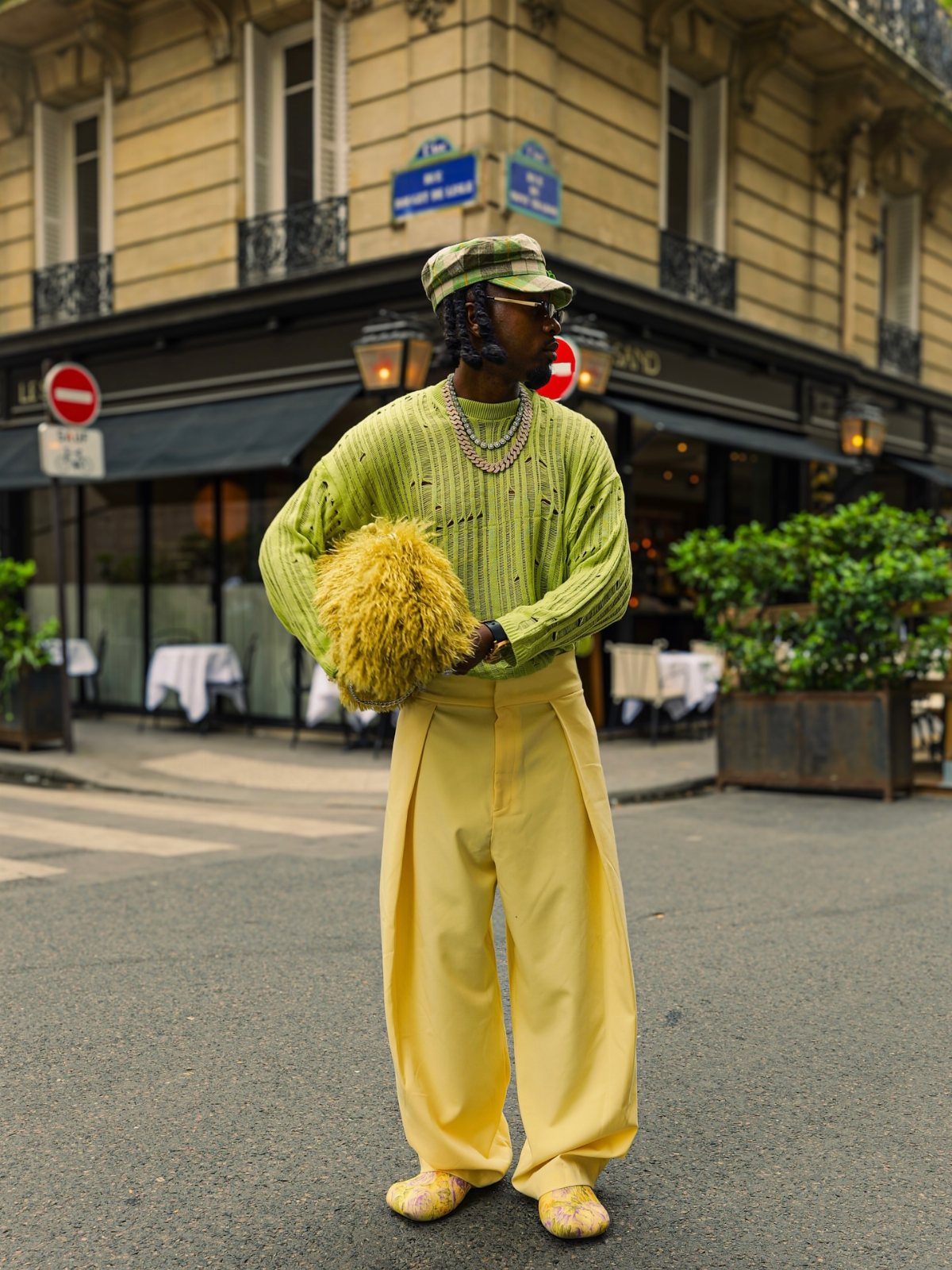 A man wearing a lime green sweater and wide yellow trousers stands on a city street.