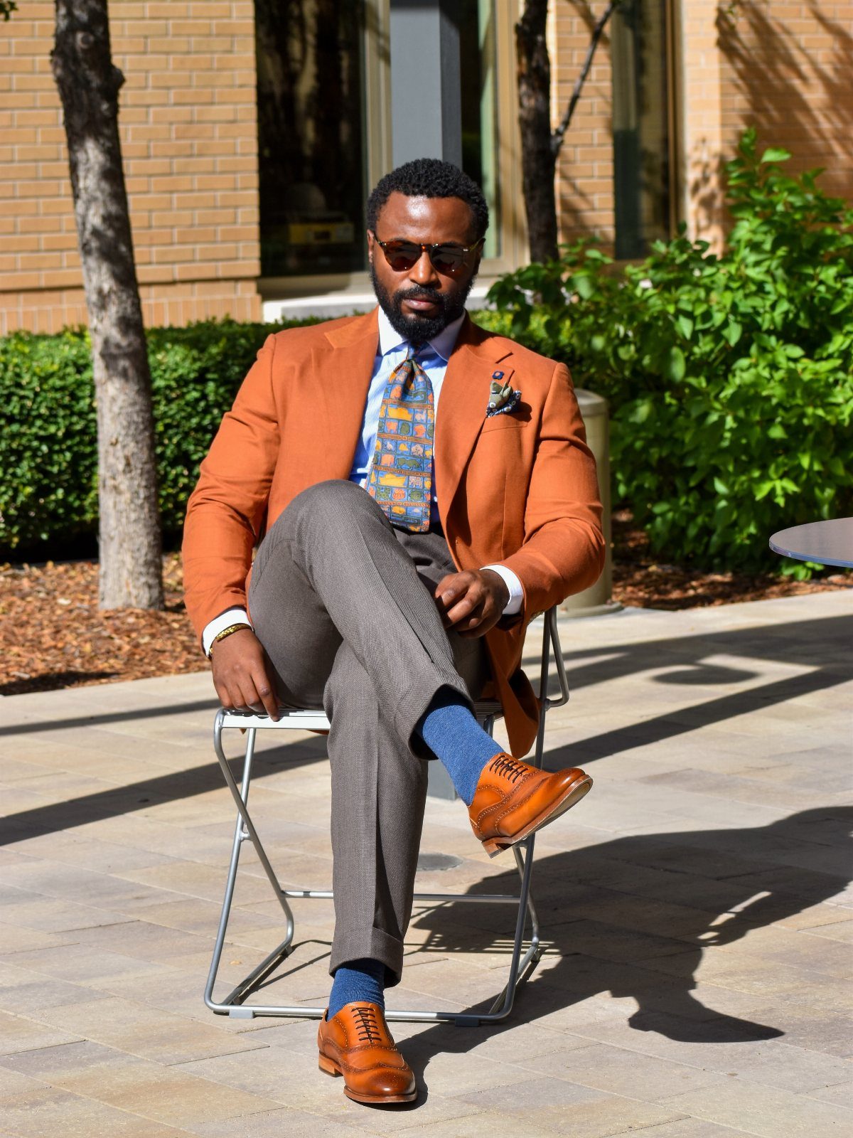 Man in an orange blazer, blue patterned tie, and grey trousers sitting on a chair outdoors.