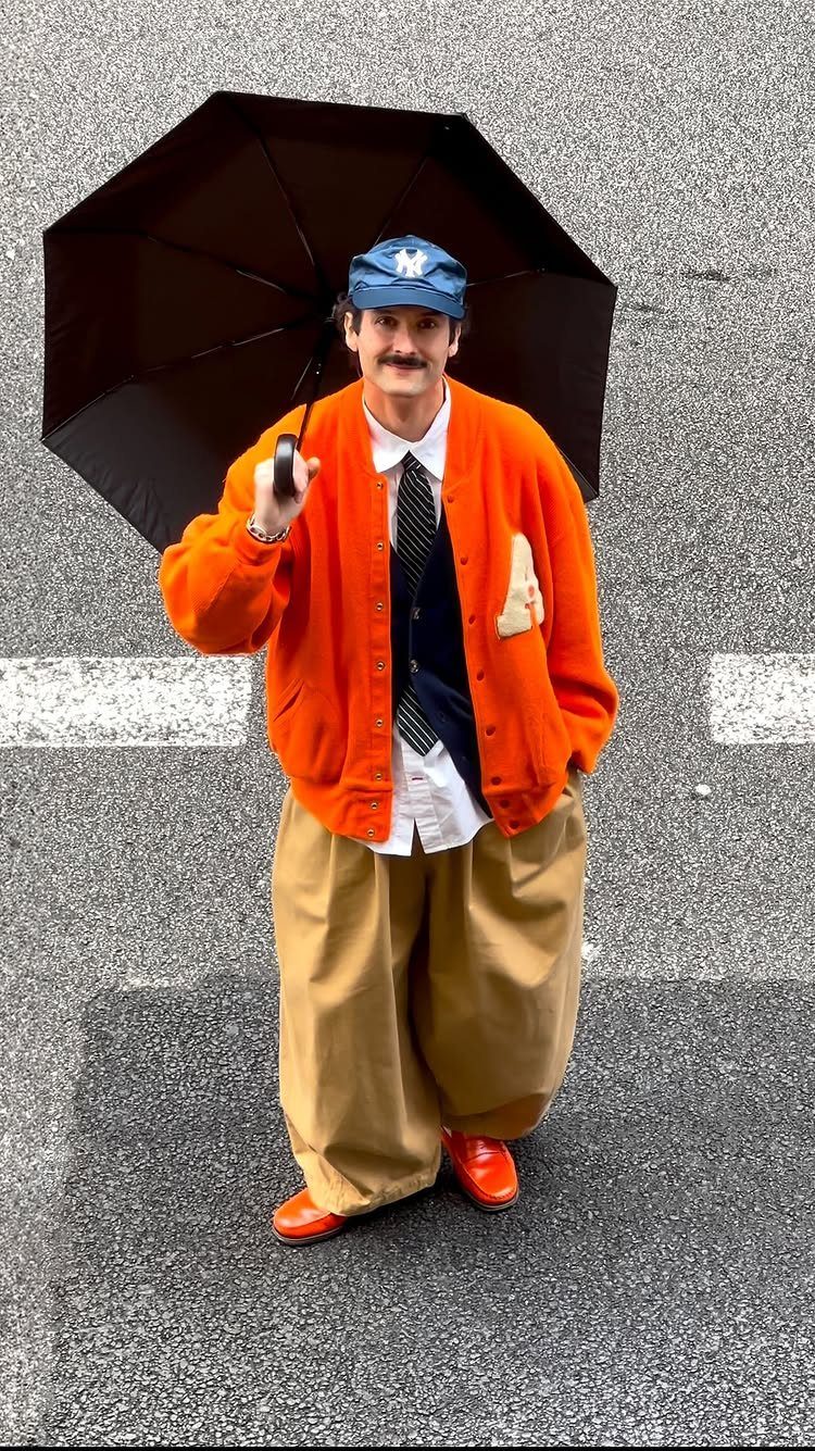 A man in a bright orange cardigan, tan trousers, and orange shoes holding a black umbrella.