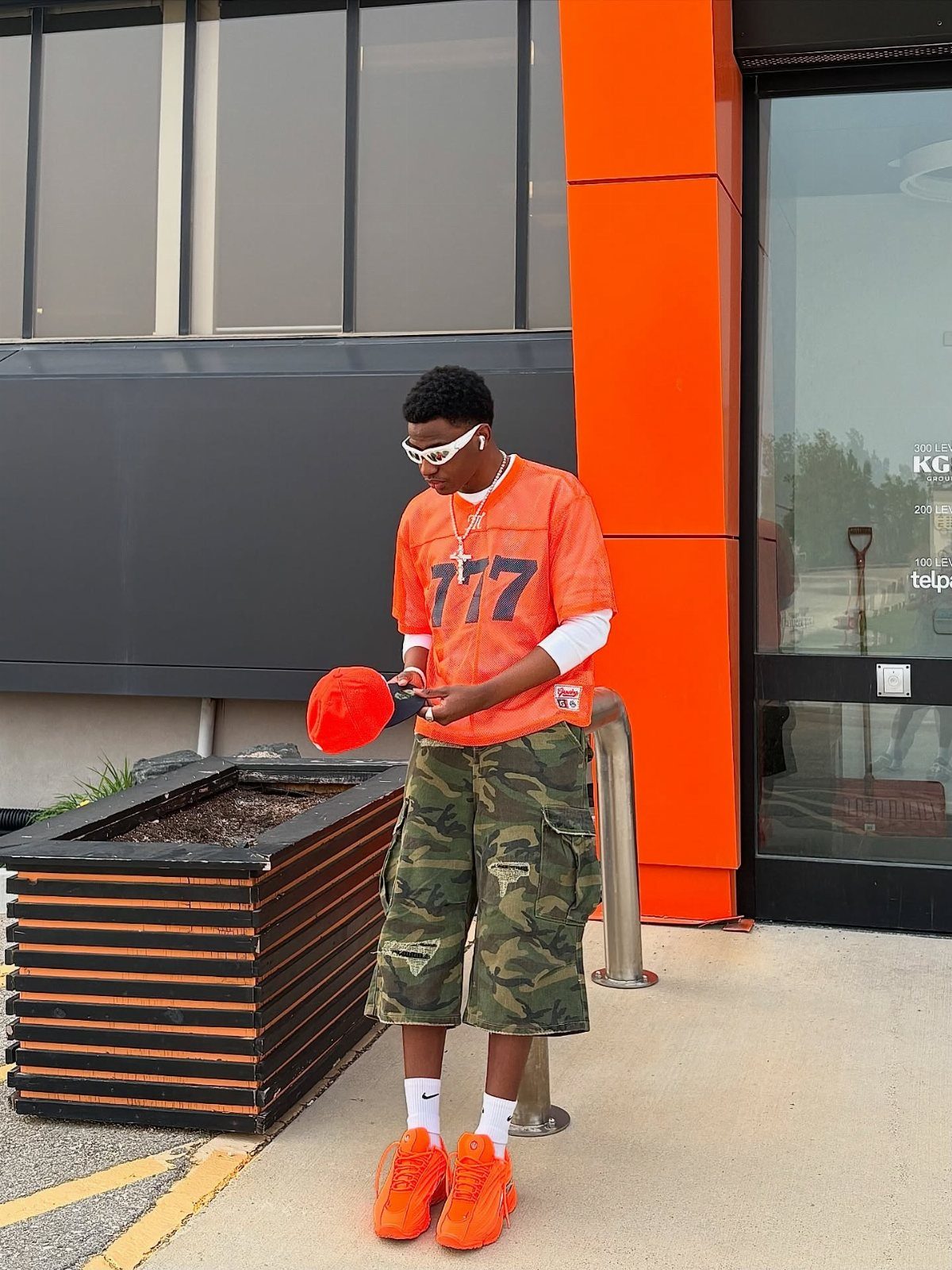 A man wearing a bright orange jersey, camouflage shorts, and orange sneakers standing outdoors.