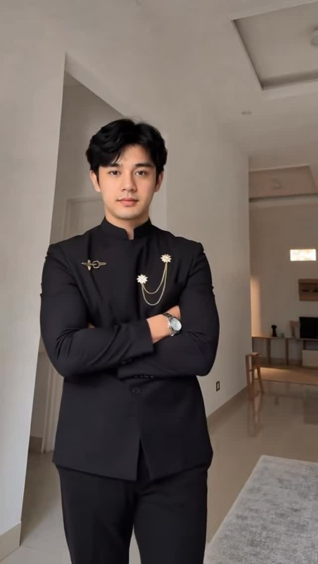 A young man wearing a black mandarin collar suit with gold floral accessories.
