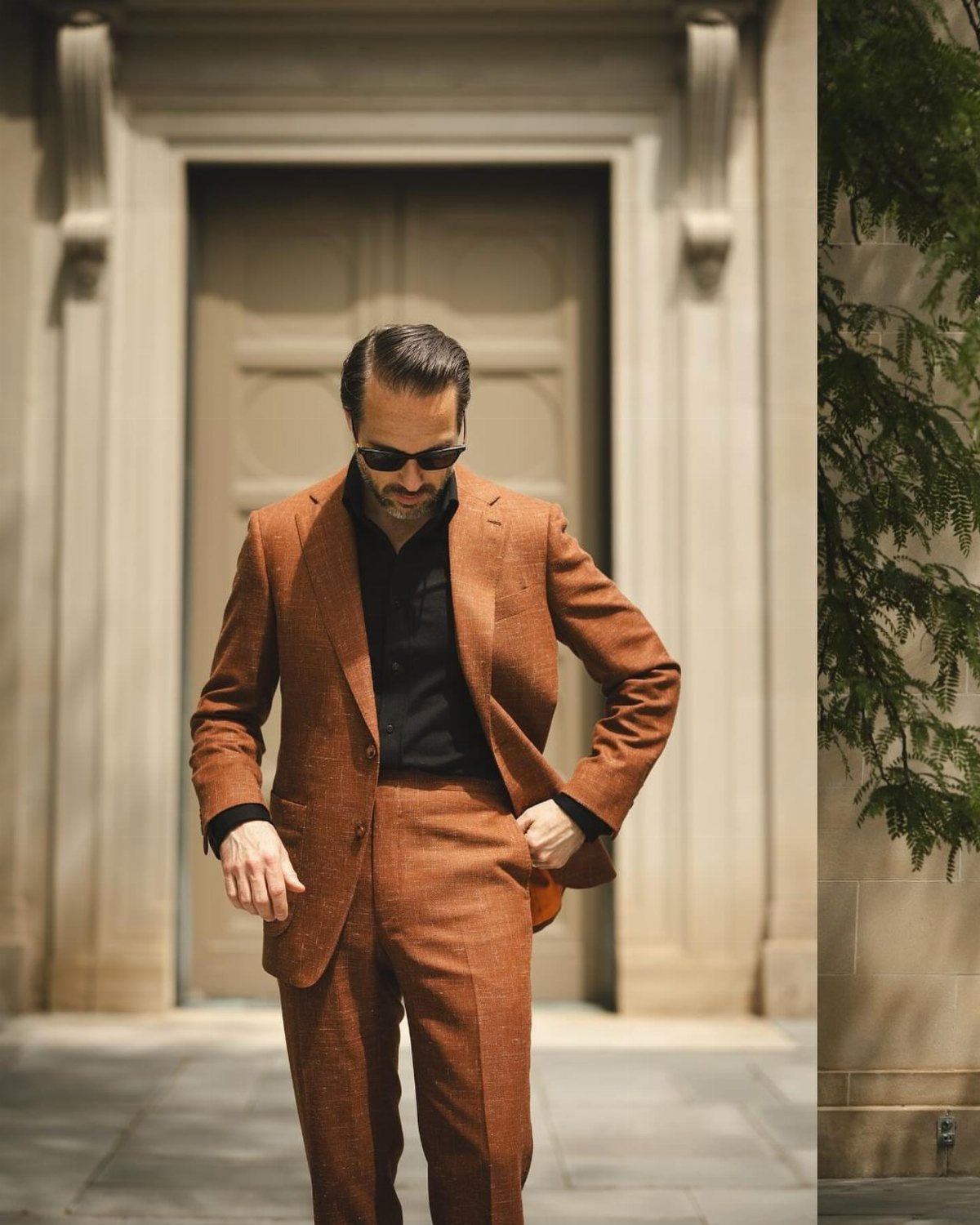 Man in a burnt orange linen suit and black shirt posing in front of a stone building.